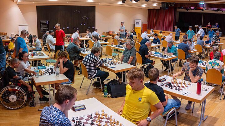 Zahlreiche Schachspieler:innen erfreuen sich im Louis Braille Saal an ihren Partien. Der Raum ist bis zum letzten Quadratmeter mit den Spieltischen und Teilnehmer:innen belegt.